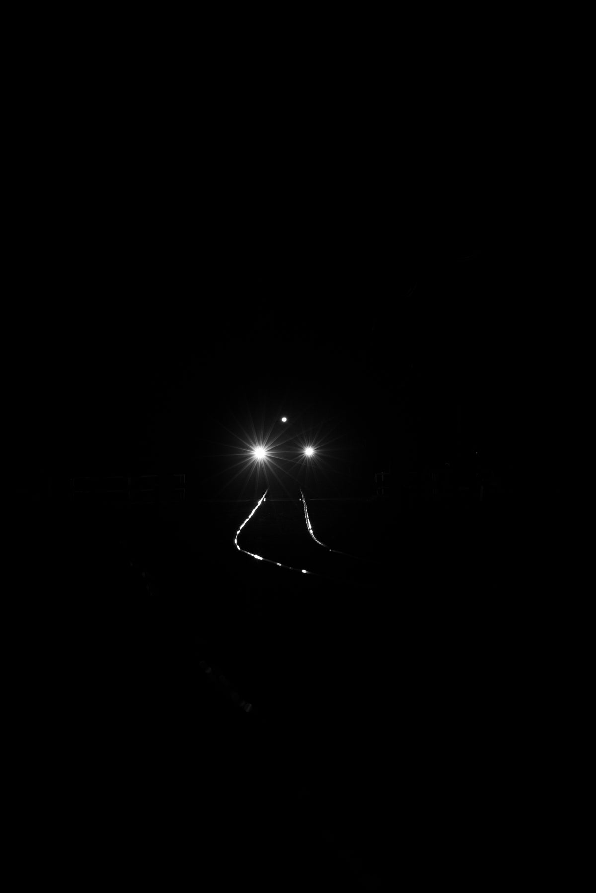 A dramatic black-and-white photograph showing the bright headlights of a vehicle on a dark road, with faint road lines visible against a pitch-black background.
