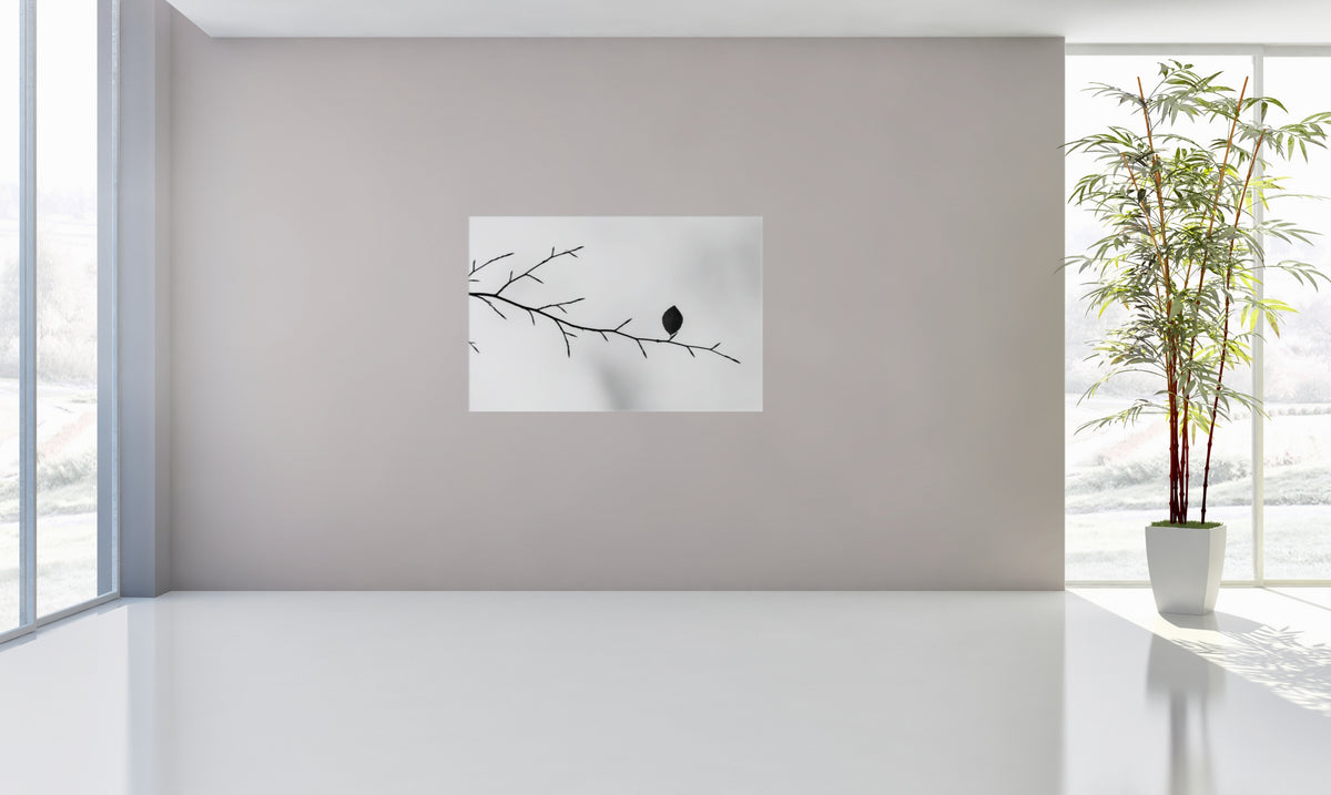 A minimalist wall art print featuring a black leaf perched on a bare branch, displayed on a light gray wall in a bright, modern room with large windows and a potted bamboo plant.