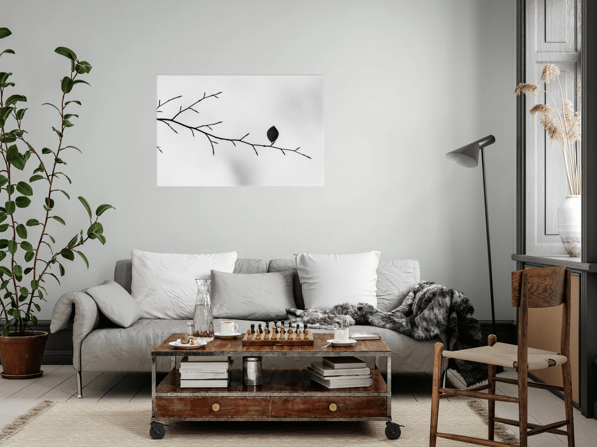A styled living room featuring a square black and white wall art print of a leaf on a branch, hung above a gray sofa with white pillows and a wooden coffee table.