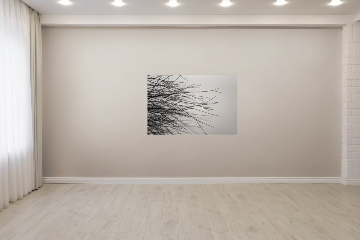 A square wall art print showing black tree branches against a light gray background, displayed on a beige wall in a modern room with light wood floors and white ceiling lights.
