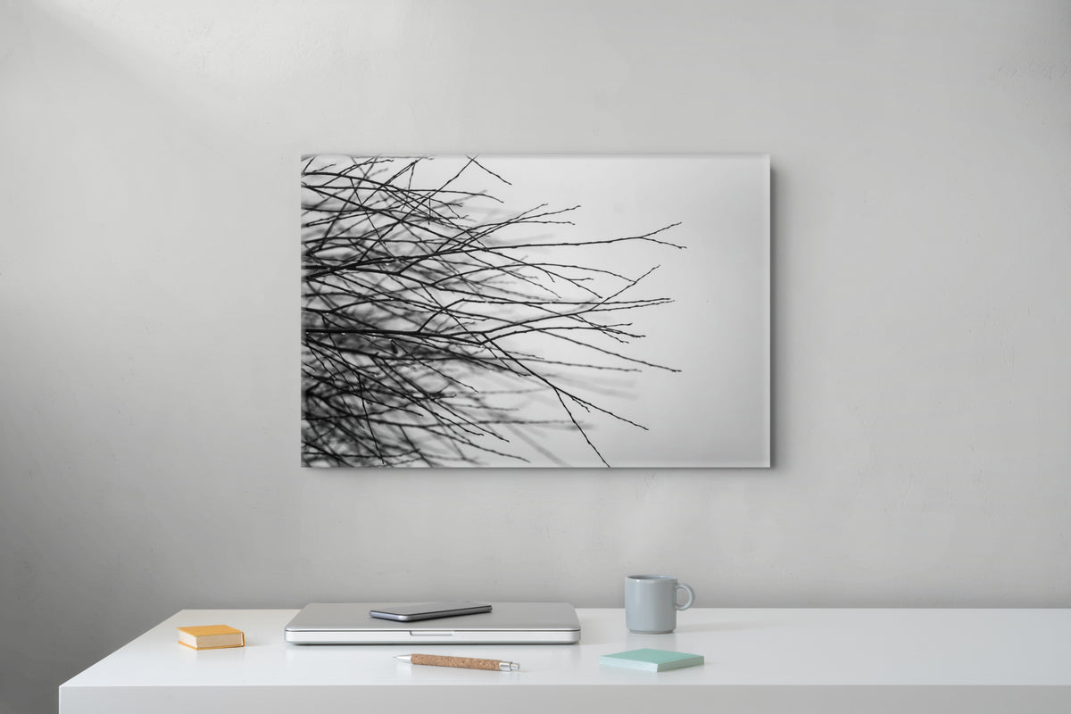 A rectangular wall art print showing black tree branches against a white background, displayed on a light gray wall above a white desk with a laptop, mug, and small notebook.