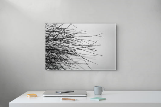 A rectangular wall art print showing black tree branches against a white background, displayed on a light gray wall above a white desk with a laptop, mug, and small notebook.