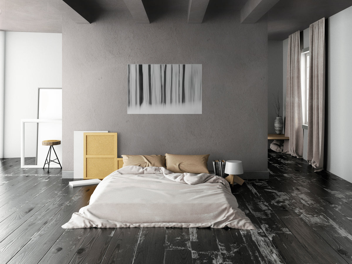 A modern minimalist bedroom featuring a low-profile platform bed with beige bedding, set against a textured gray wall with a vertical abstract art print. The room has dark wood floors, a yellow side cabinet, and a window with beige curtains on the right.