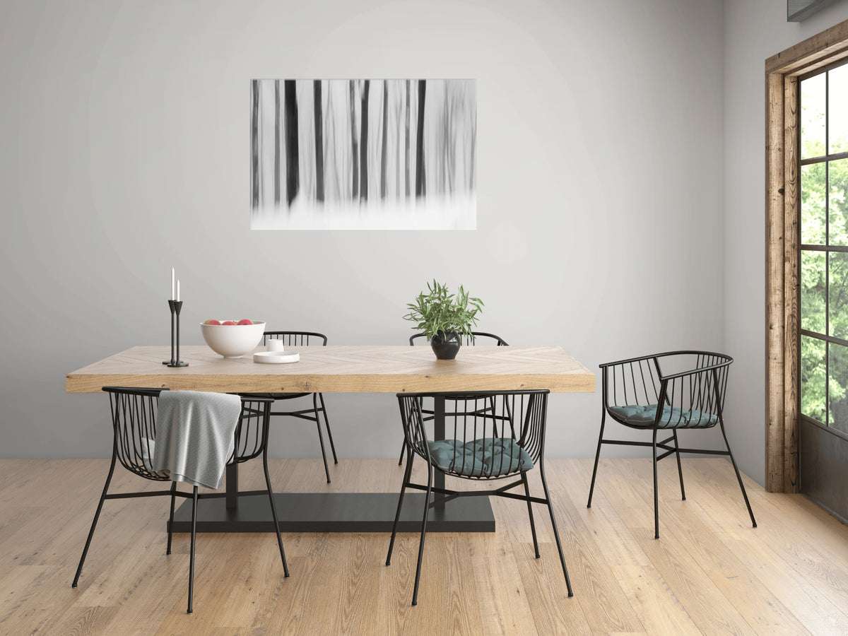 A modern dining room setup featuring a rectangular light wood table with a black metal base, surrounded by black wire-frame chairs with green cushions. The room has light wood floors, a gray wall, and a large black and white abstract canvas print hanging on the wall.