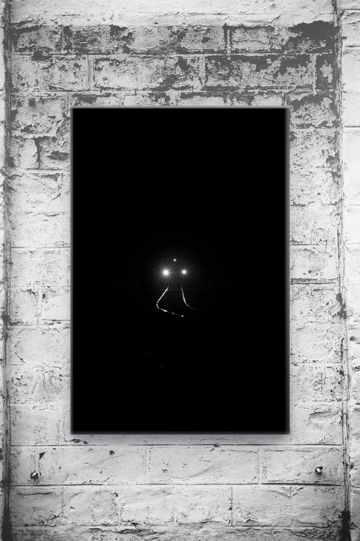 A vertical black canvas art print featuring a minimalist view of train headlights at night, mounted on a textured white brick wall.