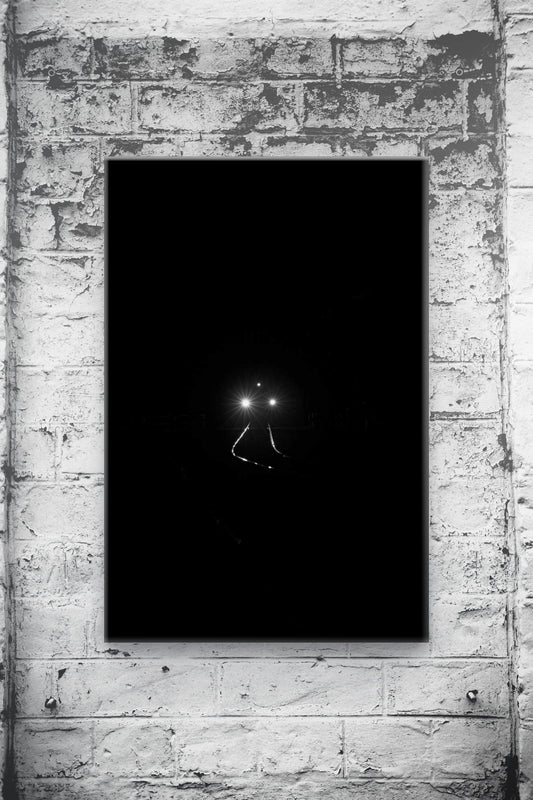A vertical black canvas art print featuring a minimalist view of train headlights at night, mounted on a textured white brick wall.