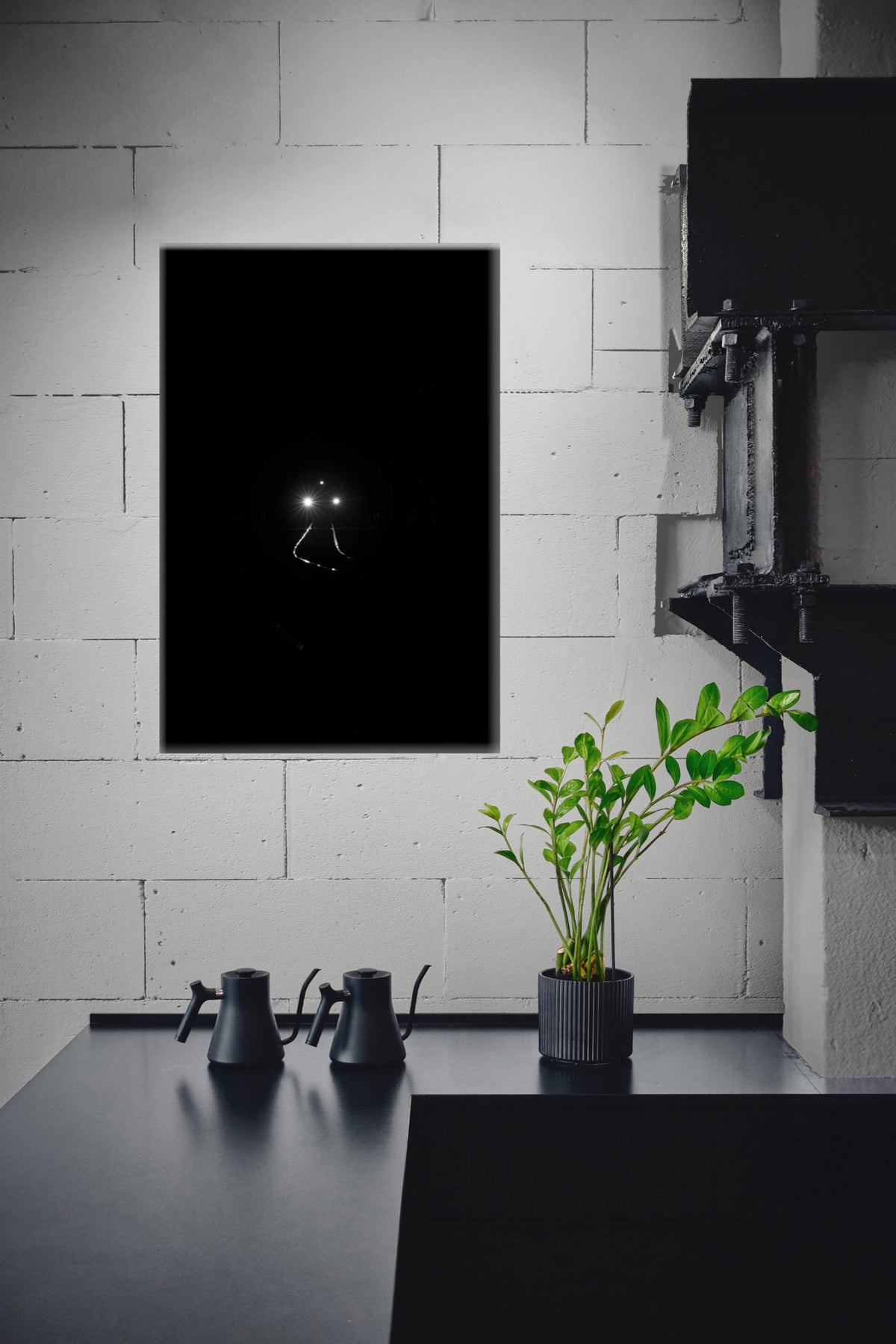 A minimalist black wall art print featuring a small glowing figure in the center, displayed on a white brick wall above a dark table with a potted green plant and two black gooseneck kettles.