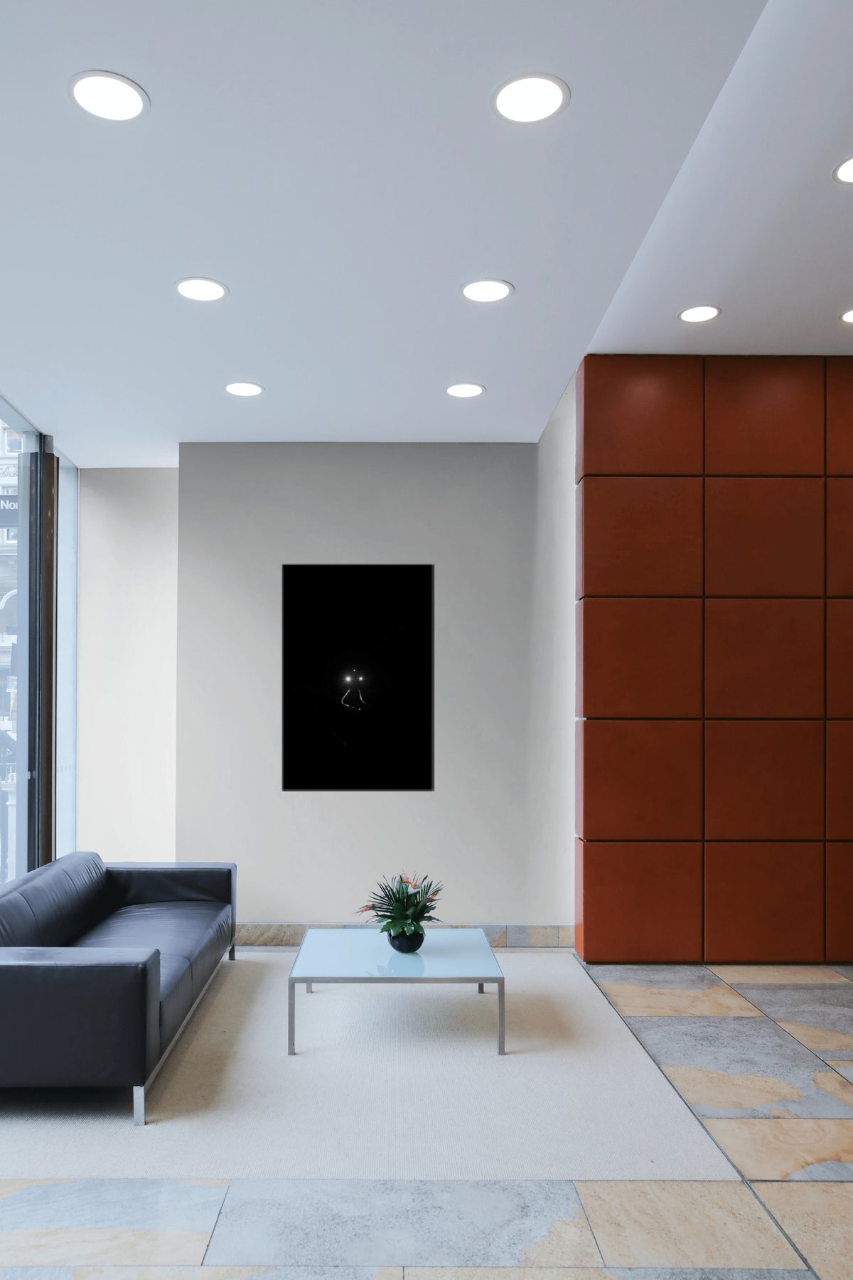 A modern interior space with a black rectangular wall art piece centered on a light gray wall, above a small white coffee table. The room includes a dark gray sofa on the left, a large rust-brown paneled wall on the right, recessed ceiling lights, and stone tile flooring.