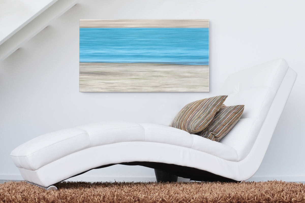 A horizontal canvas wall art print depicting a blue ocean and sandy beach, styled in a modern living room setting above a white leather chaise lounge with striped pillows.