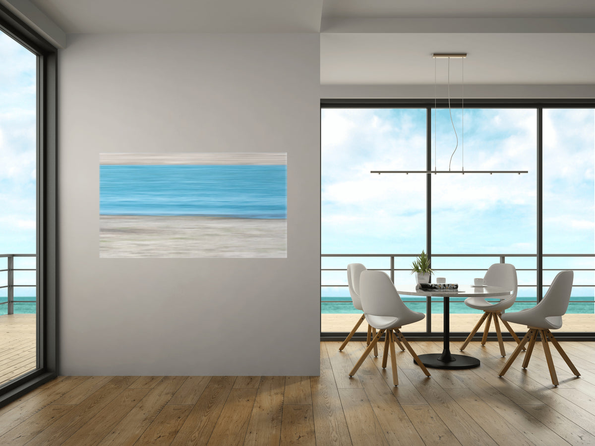 A horizontal landscape art print depicting a blue ocean and sandy beach, displayed on a light gray wall in a modern, sunlit dining room with a table, chairs, and large windows overlooking the sea.