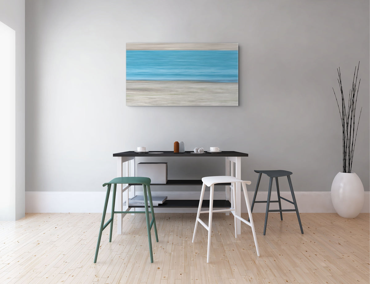 A minimalist interior scene with a framed horizontal beach-style painting on a light gray wall above a black table and three stools in green, white, and dark gray. A tall white vase with dried stems sits on a light wood floor.